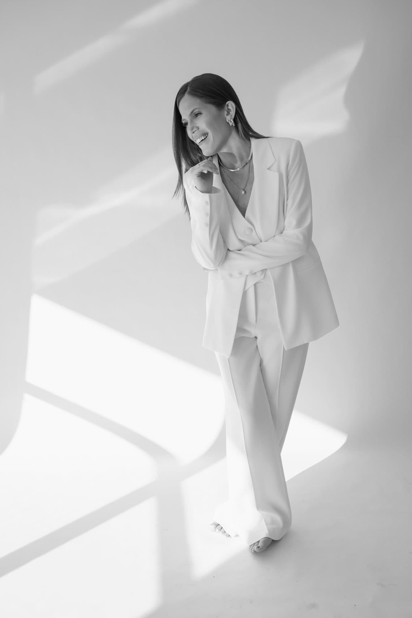 Nina Hayashi de terno claro, sorrindo, com a mão próxima ao queixo, em estúdio